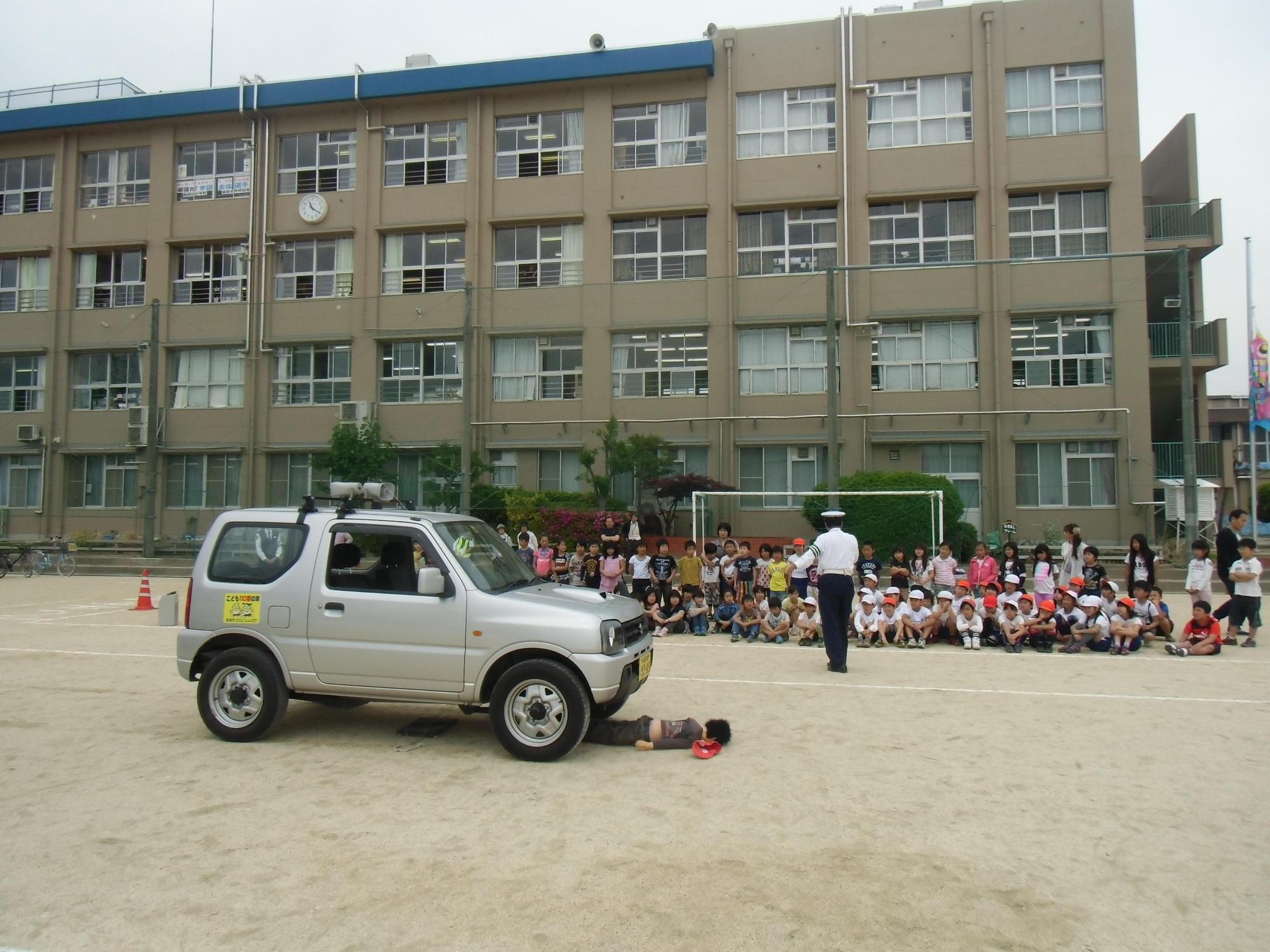 校庭で児童と同じぐらいの背丈のお人形と自動車の衝突実験を見学している写真